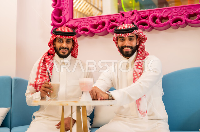 قضاء وقت ممتع برفقة الأصدقاء، اجتماع الرفاق على تناول المشروبات الباردة، جلسة وناسة في عطلة نهاية الاسبوع, النظر إلى الكاميرا بايماءات السعادة والسرور، صديقان عربيان خليجيان سعوديان يرتديان الزي التقليدي يجلسان على الأريكة في إحدى المقاهي الراقية