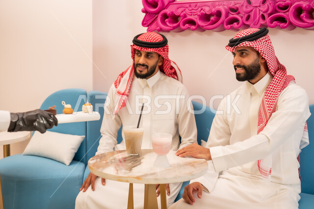 جلسة وناسة في عطلة نهاية الاسبوع, صورة مقربة لأيدي النادل يقوم بتقديم الحلويات، قضاء وقت ممتع برفقة الأصدقاء، صديقان عربيان خليجيان سعوديان يرتديان الزي التقليدي يجلسان على الأريكة في إحدى المقاهي الراقية يتناولان المشروبات الباردة
