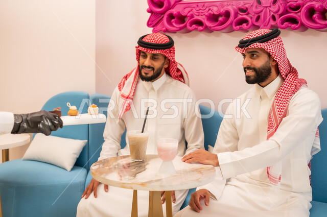 الاهتمام والعناية بالزبائن، جلسة وناسة في عطلة نهاية الاسبوع, صورة مقربة لأيدي النادل يقوم بتقديم الحلويات، قضاء وقت ممتع برفقة الأصدقاء، صديقان عربيان خليجيان سعوديان يرتديان الزي التقليدي يجلسان على الأريكة في إحدى المقاهي الراقية يتناولان المشروبات الباردة