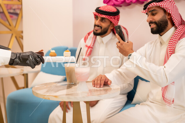 التقاط الصور التذكارية عبر الهاتف المحمول، صورة مقربة لأيدي النادل يقوم بتقديم الحلويات، قضاء وقت ممتع برفقة الأصدقاء، صديقان عربيان خليجيان سعوديان يرتديان الزي التقليدي يجلسان على الأريكة في إحدى المقاهي الراقية يتناولان المشروبات الباردة