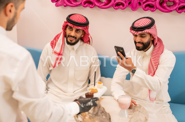 قضاء وقت ممتع برفقة الأصدقاء خارج المنزل، تناول السكريات والمشروبات الباردة، التقاط الصور التذكارية عبر الهاتف المحمول، صورة مقربة من الخلف لنادل يقوم بتقديم الحلويات، صديقان عربيان خليجيان سعوديان يرتديان الزي التقليدي يجلسان على الأريكة في إحدى المقاهي الراقية