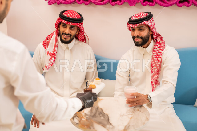 الاستمتاع برفقة الأصدقاء خارج المنزل، تناول السكريات والمشروبات الباردة، التقاط الصور التذكارية عبر الهاتف المحمول، صورة مقربة من الخلف لنادل يقوم بتقديم الحلويات، صديقان عربيان خليجيان سعوديان يرتديان الزي التقليدي يجلسان على الأريكة في إحدى المقاهي الراقية