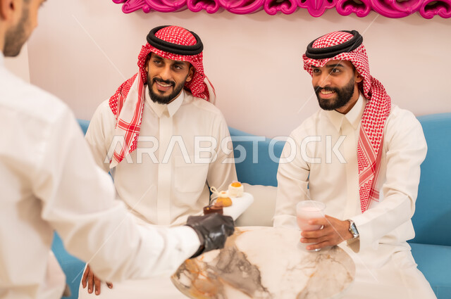 Enjoying the company of friends outside the home, eating sweets and cold drinks, taking souvenir photos via mobile phone, a close-up photo from the back of a waiter serving desserts, two Saudi Arabian Gulf friends wearing traditional dress sitting on the couch in an upscale cafe