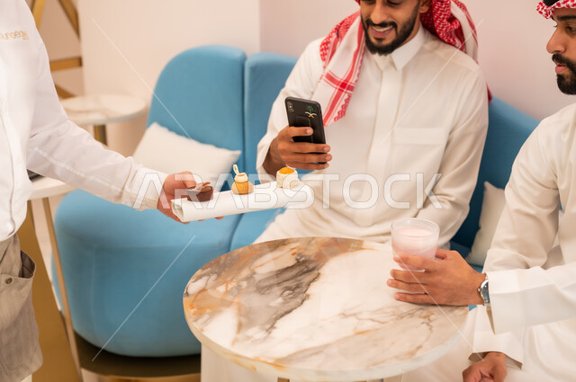 تناول السكريات والمشروبات الباردة، التقاط الصور التذكارية عبر الهاتف المحمول، صورة مقربة ليد نادل يقوم بتقديم الحلويات، قضاء وقت ممتع برفقة الأصدقاء خارج المنزل، صديقان عربيان خليجيان سعوديان يرتديان الزي التقليدي يجلسان على الأريكة في إحدى المقاهي الراقية