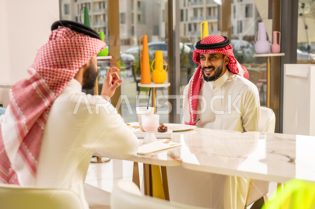 جلسة وناسة وتعارف، قضاء وقت ممتع في الخارج، شابان عربيان خليجيان سعوديان يرتديان الثوب والشماغ السعودي يجلسان في إحدى المقاهي الراقية بايماءات السعادة والسرور، تبادل الاحاديث بين الأصدقاء، مشاركة المشروبات الباردة والحلويات، تطوير العلاقات الاجتماعية