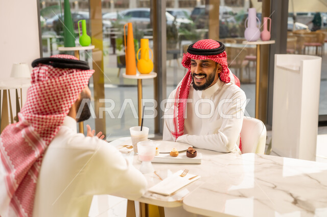 قضاء وقت ممتع في الخارج، صديقان عربيان خليجيان سعوديان يرتديان الثوب والشماغ السعودي يجلسان في إحدى المقاهي الراقية بايماءات السعادة والسرور، تبادل الاحاديث بين الأصدقاء، مشاركة المشروبات الباردة والحلويات