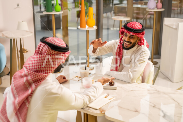 Friendships, acquaintances, and building social relationships, spending a pleasant time in an upscale café, concluding new successful business deals between businessmen, partnership in business and projects, two Saudi Arabian Gulf men wearing traditional clothing shaking hands with gestures of happiness and pleasure