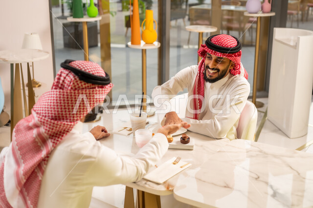 Partnership in business and projects, friendships, acquaintances and building social relationships, spending a pleasant time in an upscale cafe, concluding new successful business deals between businessmen, two Saudi Arabian Gulf men wearing traditional clothing shaking hands with gestures of happiness and pleasure