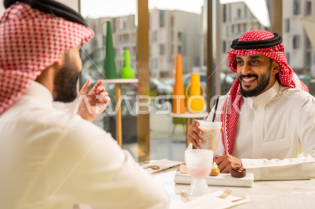 جلسة وناسة وتعارف، قضاء وقت ممتع في الخارج، شابان عربيان خليجيان سعوديان يرتديان الثوب والشماغ السعودي يجلسان في إحدى المقاهي الراقية بايماءات السعادة والسرور، تبادل الاحاديث بين الأصدقاء، مشاركة المشروبات الباردة والحلويات، تطوير العلاقات الاجتماعية
