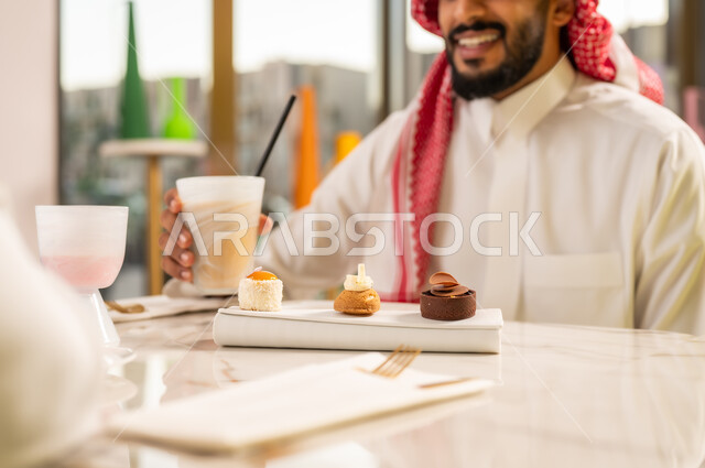 تناول الكوكتيل البارد والحلوى، محاولة التخفيف من حرارة الصيف بالمشروب المنعش، المتعة والتسلية والترفيه في الخارج، صورة مقربة لرجل عربي خليجي سعودي مبتسم يرتدي الثوب والشماغ و يجلس في مقهى راقي ويستمتع بشرب السموذي الطبيعي بإيماءات السرور والسعادة