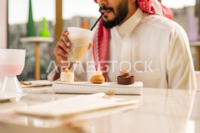 محاولة التخفيف من حرارة الصيف بالمشروب المنعش، تناول الكوكتيل البارد والحلوى، المتعة والتسلية والترفيه في الخارج، صورة مقربة لرجل عربي خليجي سعودي مبتسم يرتدي الثوب والشماغ و يجلس في مقهى راقي ويستمتع بشرب السموذي الطبيعي