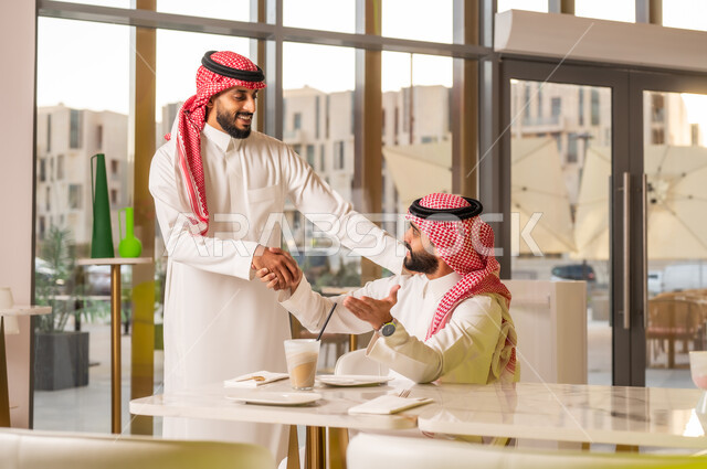 إيماءات السرور والسعادة برؤية الأصدقاء، إلقاء التحيه والسلام, رجلان عربيان خليجيان سعوديان يتقابلان في المقهى ويقومان بمصافحة بعضهما, مفهوم الاستقبال والترحيب، صداقة وتعارف وبناء علاقات اجتماعية، قضاء وقت ممتع في الخارج