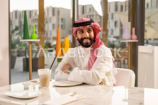 كوفي شوب راقي في المملكة العربية السعودية، محاولة التخفيف من حرارة الصيف بالمشروب المنعش، تناول السموذي البارد والحلوى، المتعة والتسلية والترفيه في الخارج، رجل عربي خليجي سعودي مبتسم يرتدي الثوب والشماغ و يجلس في المقهى وينظر إلى شيء ما بتعابير السرور والسعادة