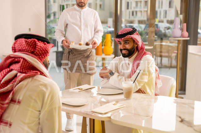 نادل يقوم بتقديم الحلويات، قضاء وقت ممتع في الخارج، صداقات وتعارف وبناء علاقات اجتماعية، الاهتمام والعناية بالزبائن، جلسة وناسة في عطلة نهاية الاسبوع, صديقان عربيان خليجيان سعوديان يرتديان الزي التقليدي يجلسان في إحدى المقاهي الراقية
