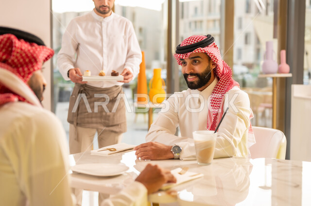 قضاء وقت ممتع في الخارج، صداقات وتعارف وبناء علاقات اجتماعية، الاهتمام والعناية بالزبائن، جلسة وناسة في عطلة نهاية الاسبوع, نادل يقوم بتقديم الحلويات، صديقان عربيان خليجيان سعوديان يرتديان الزي التقليدي يجلسان في إحدى المقاهي الراقية
