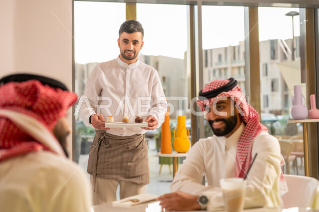 قضاء وقت ممتع في الخارج، صداقات وتعارف وبناء علاقات اجتماعية، الاهتمام والعناية بالزبائن، جلسة وناسة في عطلة نهاية الاسبوع, نادل يقوم بتقديم الحلويات، صديقان عربيان خليجيان سعوديان يرتديان الزي التقليدي يجلسان في إحدى المقاهي الراقية