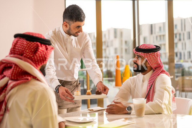 نادل يقوم بتقديم الحلويات، قضاء وقت ممتع في الخارج، صداقات وتعارف وبناء علاقات اجتماعية، الاهتمام والعناية بالزبائن، جلسة وناسة في عطلة نهاية الاسبوع, صديقان عربيان خليجيان سعوديان يرتديان الزي التقليدي يجلسان في إحدى المقاهي الراقية
