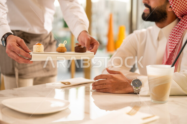 العناية والاهتمام بالزبون، نادل يقوم بتقديم الحلا، كوفي شوب راقي في المملكة العربية السعودية، تناول السموذي البارد والحلوى، المتعة والتسلية والترفيه في الخارج، صورة مقربة لرجل عربي خليجي سعودي مبتسم يرتدي الثوب والشماغ يجلس في المقهى بتعابير السرور والسعادة