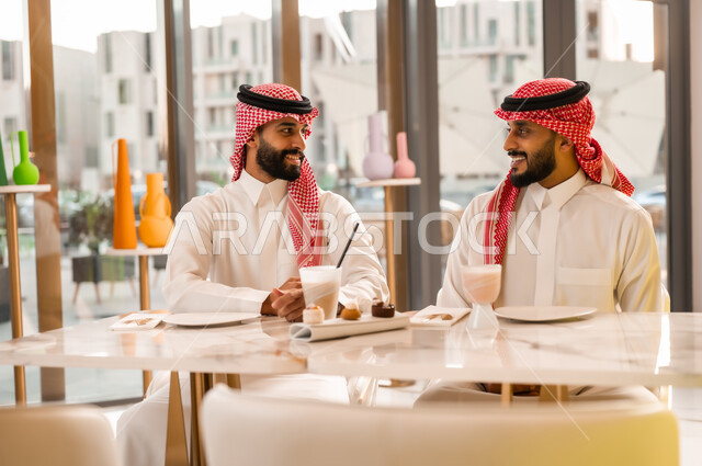جلسة وناسة وتعارف، قضاء وقت ممتع في الخارج، شابان عربيان خليجيان سعوديان يرتديان الثوب والشماغ السعودي يجلسان في إحدى المقاهي الراقية بايماءات السعادة والسرور، تبادل الاحاديث بين الأصدقاء، مشاركة المشروبات الباردة والحلويات، تطوير العلاقات الاجتماعية