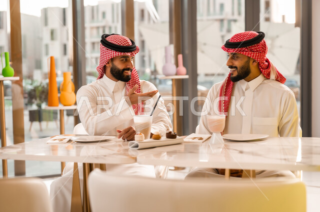 الإشارة إلى شيء ما، جلسة تسلية وترفيه شبابية، تبادل أطراف الحديث، صداقة وتعارف وبناء علاقات اجتماعية، ايماءات السعادة والسرور، شابان عربيان خليجيان سعوديان يرتديان الثوب والشماغ يجلسان في المقهى، قضاء وقت ممتع في الخارج، تناول المشروب البارد والحلوى