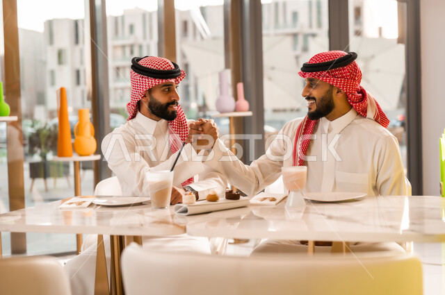 Ease of automated payment in Saudi cafes, a man paying the bill via mobile phone, using modern technical means in electronic payment, having a cold drink and dessert, entertainment and fun with friends, two Saudi Arabian Gulf men wearing traditional dress sitting in a cafe