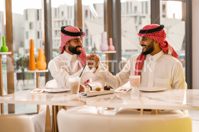 عقد صفقات عمل جديدة ناجحة، تبادل الاحاديث بين الأصدقاء، مشاركة المشروب البارد والحلوى، صداقة وتعارف وبناء علاقات اجتماعية، قضاء وقت ممتع في المقهى، رجلان عربيان خليجيان سعوديان يرتديان الزي التقليدي يقومان بمصافحة بعضهما بايماءات السعادة والسرور