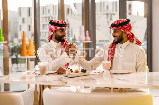 تبادل الاحاديث بين الأصدقاء، مشاركة المشروب البارد والحلوى، صداقة وتعارف وبناء علاقات اجتماعية، قضاء وقت ممتع في المقهى، عقد صفقات عمل جديدة ناجحة، رجلان عربيان خليجيان سعوديان يرتديان الزي التقليدي يقومان بمصافحة بعضهما بايماءات السعادة والسرور