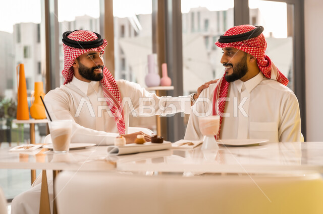 تبادل أطراف الحديث، صداقة وتعارف وبناء علاقات اجتماعية، ايماءات المحبة والسرور، شابان عربيان خليجيان سعوديان يرتديان الثوب والشماغ يجلسان في المقهى، قضاء وقت ممتع في الخارج، جلسة تسلية وترفيه شبابية
