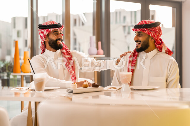 جلسة تسلية وترفيه شبابية، تبادل أطراف الحديث، صداقة وتعارف وبناء علاقات اجتماعية، ايماءات المحبة والسرور، شابان عربيان خليجيان سعوديان يرتديان الثوب والشماغ يجلسان في المقهى، قضاء وقت ممتع في الخارج