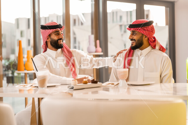 تبادل أطراف الحديث، صداقة وتعارف وبناء علاقات اجتماعية، ايماءات المحبة والسرور، شابان عربيان خليجيان سعوديان يرتديان الثوب والشماغ يجلسان في المقهى، قضاء وقت ممتع في الخارج، جلسة تسلية وترفيه شبابية