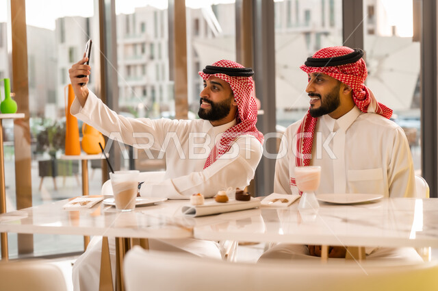 التقاط صورة سيلفي تذكارية باستخدام الهاتف المحمول بإيماءات السعادة والسرور، تناول المشروب البارد والمنعش والحلوى،  التسلية والترفيه والمتعه مع الأصدقاء، رجلان عربيان خليجيان سعوديان يرتديان الزي التقليدي السعودي يجلسان في المقهى