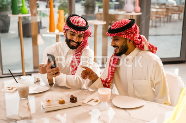 تصفح مواقع التواصل الاجتماعي، التسلية والترفيه والمتعه مع الأصدقاء، رجلان عربيان خليجيان سعوديان يرتديان الزي التقليدي السعودي يجلسان في المقهى يقومان بمشاهدة البرامج والمقاطع الترفيهية عبر الهاتف المحمول، المشاركة في تناول المشروب البارد والحلوى بتعابير السعادة