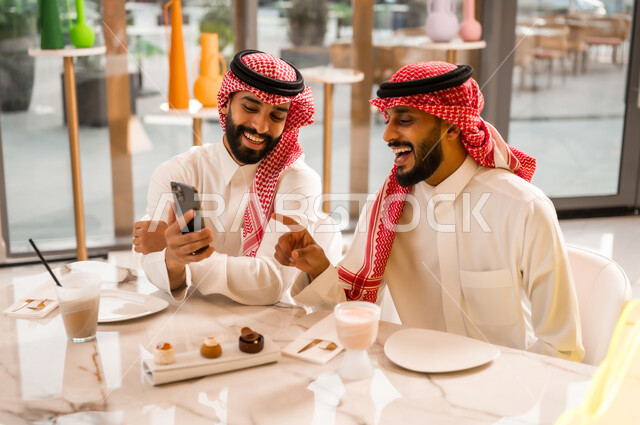 مشاهدة البرامج والمقاطع الترفيهية عبر الهاتف المحمول، التسلية والترفيه والمتعه مع الأصدقاء، رجلان عربيان خليجيان سعوديان يرتديان الزي التقليدي السعودي يجلسان في المقهى، المشاركة في تناول المشروب البارد والحلوى بتعابير السعادة والسرور