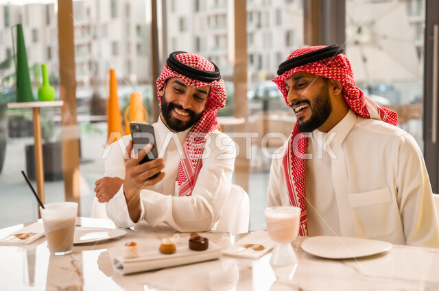 مشاهدة البرامج والمقاطع الترفيهية عبر الهاتف المحمول، التسلية والترفيه والمتعه مع الأصدقاء، رجلان عربيان خليجيان سعوديان يرتديان الزي التقليدي السعودي يجلسان في المقهى، المشاركة في تناول المشروب البارد والحلوى بتعابير السعادة والسرور