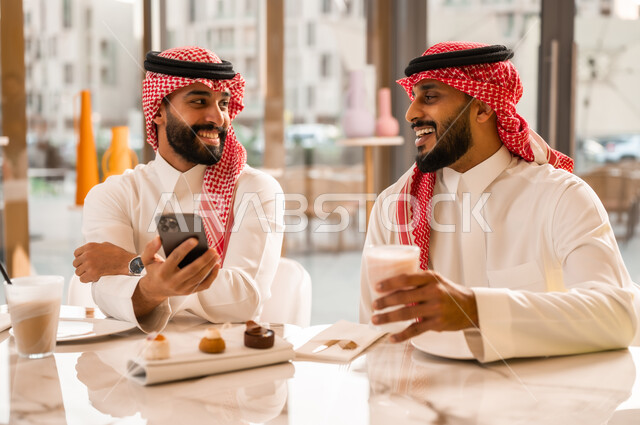 تصفح مواقع التواصل الاجتماعي، التسلية والترفيه والمتعه مع الأصدقاء، رجلان عربيان خليجيان سعوديان يرتديان الزي التقليدي السعودي يجلسان في المقهى يقومان بمشاهدة البرامج والمقاطع الترفيهية عبر الهاتف المحمول، المشاركة في تناول المشروب البارد والحلوى بتعابير السعادة