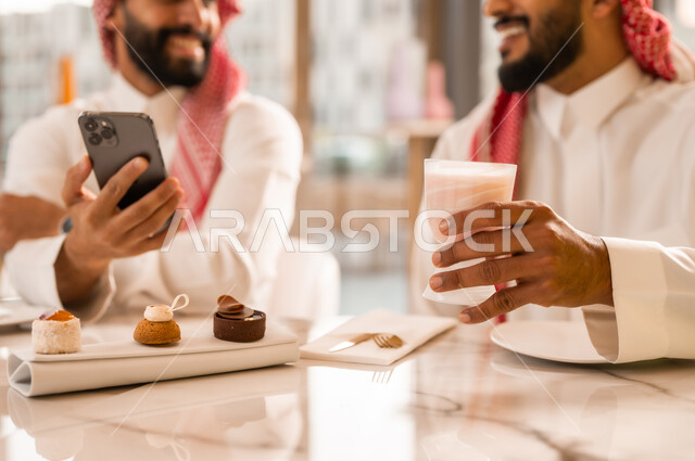 تناول المشروب البارد والحلوى بتعابير السعادة والسرور، مشاهدة البرامج والمقاطع الترفيهية عبر الهاتف المحمول، التسلية والترفيه والمتعه مع الأصدقاء، صورة مقربة لرجلين عربيين خليجيين سعوديين يرتديان الزي التقليدي السعودي يجلسان في المقهى