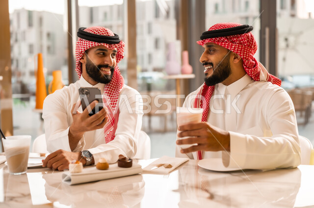 تصفح مواقع التواصل الاجتماعي، التسلية والترفيه والمتعه مع الأصدقاء، رجلان عربيان خليجيان سعوديان يرتديان الزي التقليدي السعودي يجلسان في المقهى يقومان بمشاهدة البرامج والمقاطع الترفيهية عبر الهاتف المحمول، المشاركة في تناول المشروب البارد والحلوى بتعابير السعادة