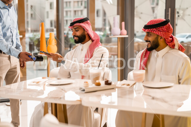 سهولة الدفع الالي في الكافيهات السعودية, رجل يقوم بدفع الحساب عبر الهاتف المحمول, استخدام الوسيلة التقنية الحديثة في الدفع الالكتروني، تناول المشروب البارد والحلوى، التسلية والمتعه مع الأصدقاء، رجلان عربيان خليجيان سعوديان يرتديان الزي التقليدي يجلسان في المقهى