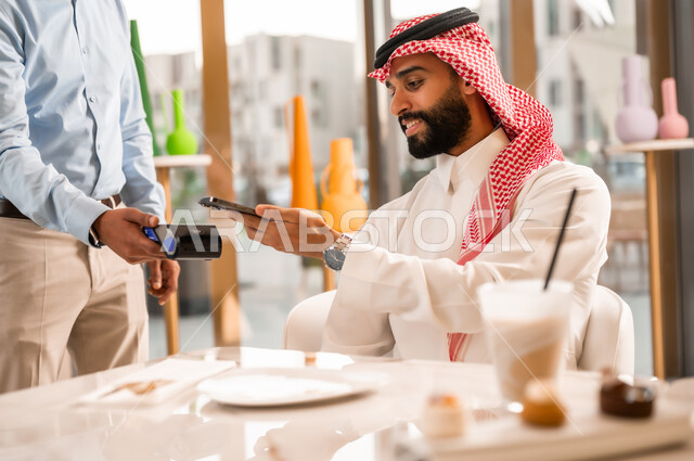 سهولة الدفع الالي في الكافيهات السعودية, رجل عربي خليجي سعودي يرتدي الثوب والشماغ يقوم بدفع الحساب عبر الهاتف المحمول, استخدام الوسيلة التقنية الحديثة في الدفع الالكتروني، تناول المشروب البارد والحلوى، التسلية والمتعه في الخارج