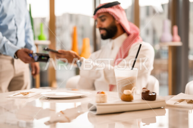 Using modern technology in electronic payment, ease of automated payment in Saudi cafes, a Saudi Arabian Gulf man wearing a thobe and shemagh paying the bill via mobile phone, drinking a cold drink and dessert, entertainment and enjoyment outside, a close-up of a plate of delicious desserts