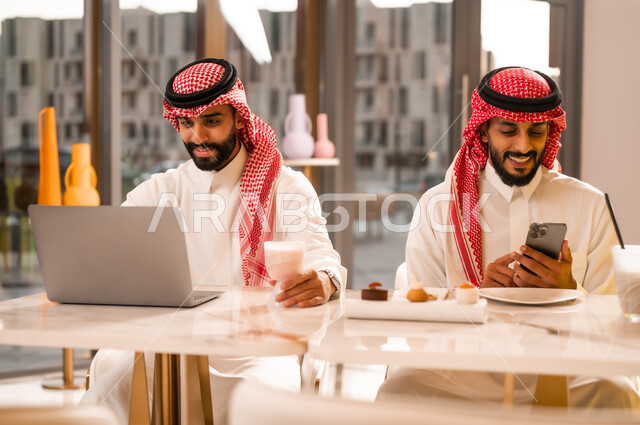 العمل عبر الحاسوب المحمول، وسيلة تقنية حديثة ، شابان عربيان خليجيان سعوديان يرتديان الثوب والشماغ يجلسان في مقهى لإنجاز المهام عن بعد، قضاء وقت ممتع في الخارج، مطعم وكافيه راقي في المملكة العربية السعودية، تصفح مواقع الانترنت عبر الهاتف