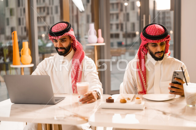 Having a cold and refreshing drink, working on a laptop, a modern technology tool, two young Saudi Arabian Gulf men wearing a thobe and a shemagh sitting in a cafe to complete tasks remotely, having a good time outside, a high-end restaurant and cafe in Saudi Arabia, browsing the internet on a phone