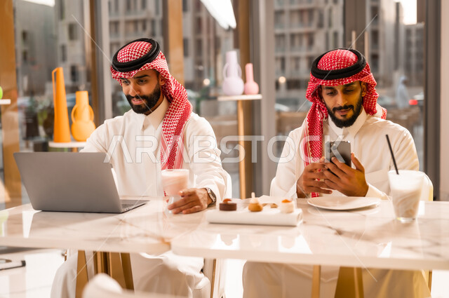 إيماءات الجدية والتركيز في العمل عبر الحاسوب المحمول، وسيلة تقنية حديثة ، شابان عربيان خليجيان سعوديان يرتديان الثوب والشماغ يجلسان في مقهى لإنجاز المهام عن بعد، قضاء وقت ممتع في الخارج، مطعم وكافيه راقي في المملكة العربية السعودية، تصفح مواقع الانترنت عبر الهاتف