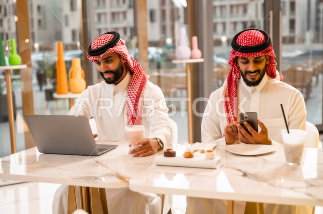 Having a cold and refreshing drink, working on a laptop, a modern technology tool, two young Saudi Arabian Gulf men wearing a thobe and a shemagh sitting in a cafe to complete tasks remotely, having a good time outside, a high-end restaurant and cafe in Saudi Arabia, browsing the internet on a phone