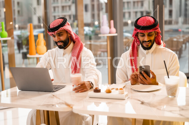 إيماءات الجدية والتركيز في العمل عبر الحاسوب المحمول، وسيلة تقنية حديثة ، شابان عربيان خليجيان سعوديان يرتديان الثوب والشماغ يجلسان في مقهى لإنجاز المهام عن بعد، قضاء وقت ممتع في الخارج، مطعم وكافيه راقي في المملكة العربية السعودية، تصفح مواقع الانترنت عبر الهاتف