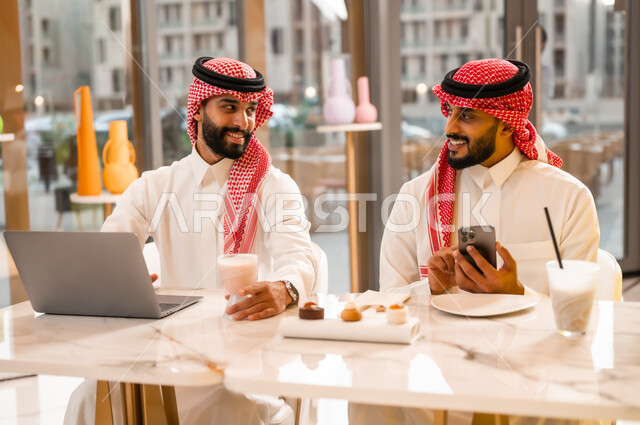 نقاش وحوار لإدارة الأعمال وتنظيمها، تناول المشروب البارد والمنعش، العمل عبر الحاسوب المحمول، وسيلة تقنية حديثة، شابان عربيان خليجيان سعوديان يرتديان الثوب والشماغ يجلسان في مقهى لإنجاز المهام عن بعد، قضاء وقت ممتع في الخارج، استخدام الهاتف الجوال