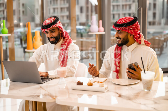 المناقشة والحوار لإدارة الأعمال وتنظيمها، الاستمتاع بتناول المشروب البارد والمنعش في الخارج، العمل عبر الحاسوب المحمول، وسيلة تقنية حديثة، شابان عربيان خليجيان سعوديان يرتديان الثوب والشماغ يجلسان في مقهى لإنجاز المهام عن بعد، استخدام الهاتف الجوال