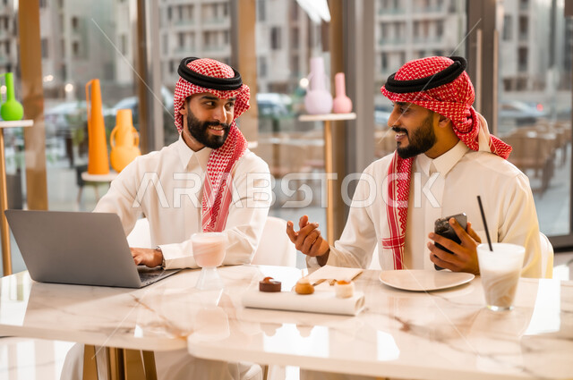 نقاش وحوار لإدارة الأعمال وتنظيمها، تناول المشروب البارد والمنعش، العمل عبر الحاسوب المحمول، وسيلة تقنية حديثة، شابان عربيان خليجيان سعوديان يرتديان الثوب والشماغ يجلسان في مقهى لإنجاز المهام عن بعد، قضاء وقت ممتع في الخارج، استخدام الهاتف الجوال