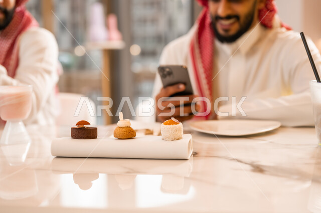 Sharing a cold drink and dessert, having a good time with friends, close-up of a plate of delicious dessert, a smiling Saudi Arabian Gulf man wearing traditional dress sitting with his friend in a cafe and browsing the internet on his mobile phone with happy gestures, using a modern and advanced technical device