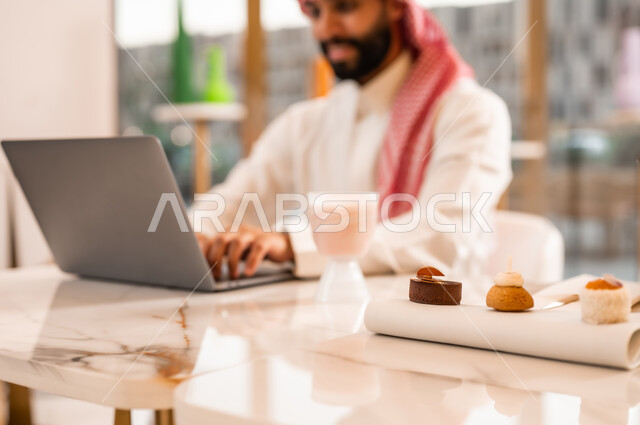 مطعم راقي في السعودية، صورة مقربة لطبق من الحلوى، استخدام الحاسوب المحمول لإنجاز الأعمال والمشاريع، الاستمتاع بالاجواء الهادئه خارج المنزل، رجل عربي خليجي سعودي يرتدي الثوب والشماغ يجلس في المقهى يقووم بالعمل عبر الانترنت، تناول الحلا والسكريات في الخارج